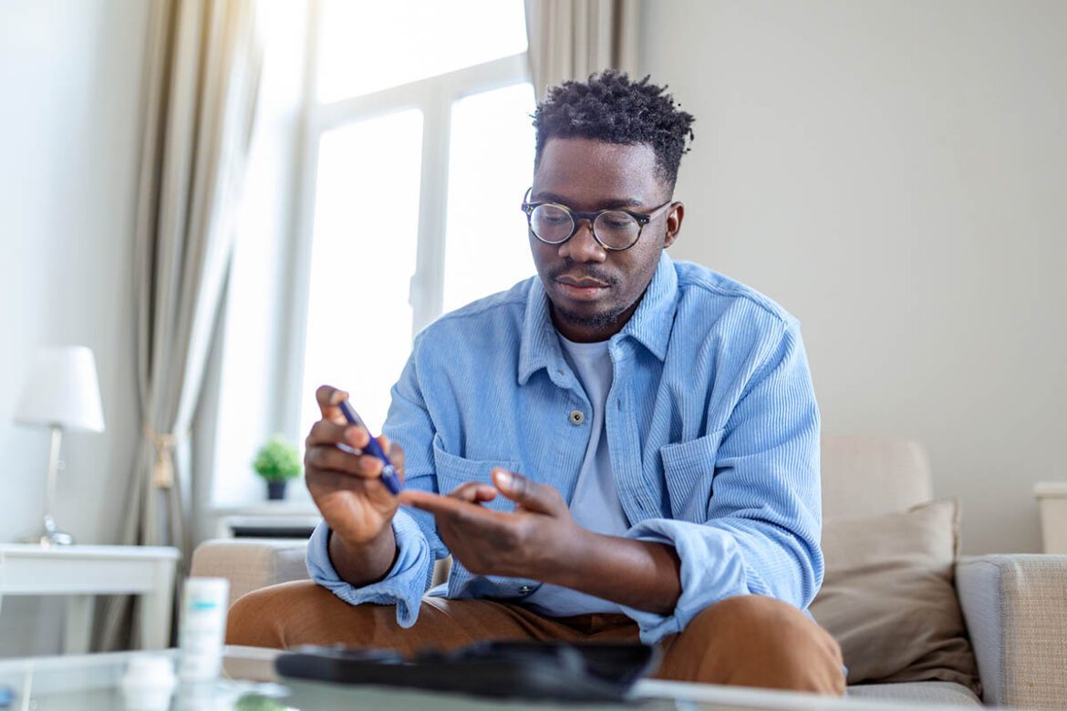African man is sitting at the sofa and taking blood from his finger due to diabetes. the daily life of a man of african american ethnicity person with a chronic illness who is using glucose tester.